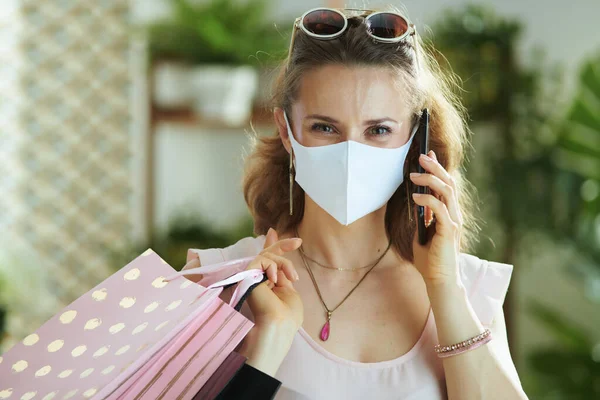コロナウイルスのパンデミックの間の生活 ピンクのブラウスを着た現代女性の買い物客の肖像と医療マスクと紙のショッピングバッグがスマートフォンで話しています — ストック写真