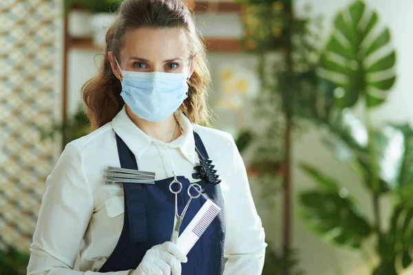 Small business survival after covid-19 pandemic. Portrait of stylish small business owner woman in apron with medical mask, gloves, hair comb and scissors.