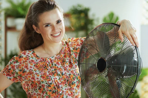 夏の暑さ 肖像画の笑顔若いです女性で花のブラウスで電気ファンで現代のリビングルームで晴れた暑い夏の日 — ストック写真