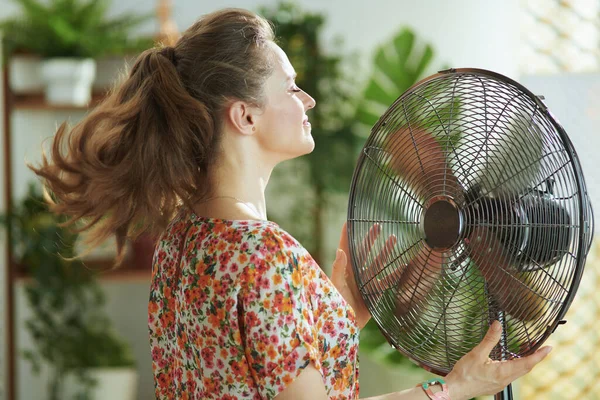 夏の暑さ 現代の笑顔40歳の女性です花のブラウスで現代のリビングルームで日当たりの良い暑い夏の日自分自身を扇 — ストック写真