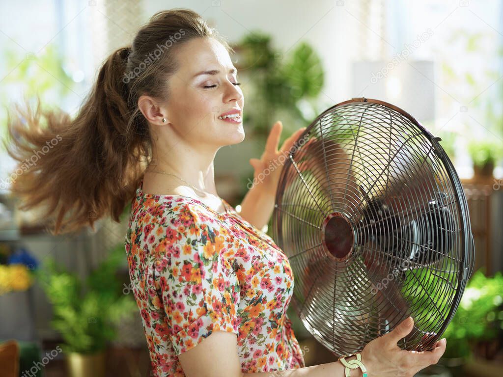 Summer heat. smiling modern woman in floral blouse in the modern house in sunny hot summer day fanning herself.