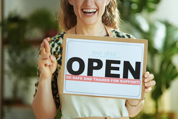 Opening small business after covid-19 pandemic. Closeup on smiling middle aged small business owner woman in apron with crossed fingers showing open sign.