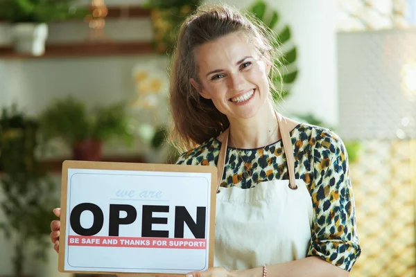 Opening small business after covid-19 pandemic. Portrait of happy modern 40 years old small business owner woman in apron showing open sign.