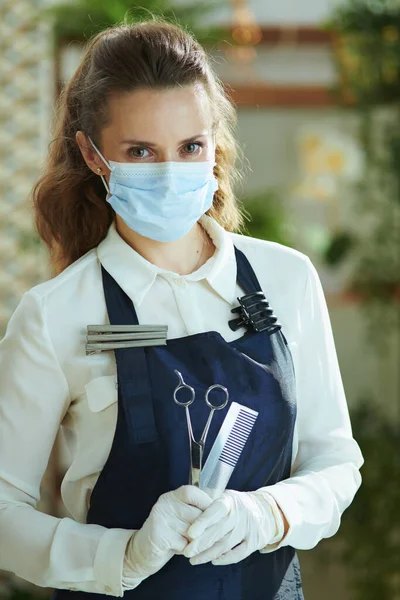 Opening small business after covid-19 pandemic. Portrait of modern small business owner woman in apron with medical mask, gloves, hair comb and scissors.