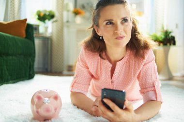 Düşünceli genç ev hanımı bluz ve kumbaralı beyaz pantalon akıllı telefondan halı temizleme servisi ararken güneşli bir günde modern evde beyaz halıda yatıyordu..