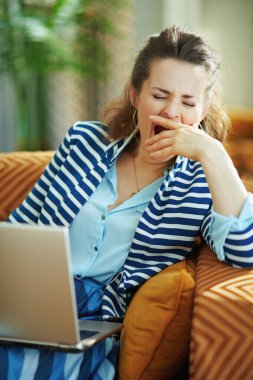 Mavi bluzlu ve çizgili ceketli yorgun genç bir kadın güneşli bir günde modern evdeki dizüstü bilgisayarla kanepede oturuyor..