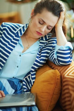 Stresli, mavi bluzlu ve çizgili ceketli modern kadın güneşli bir günde modern oturma odasında dizüstü bilgisayarla divanda uyuyor..