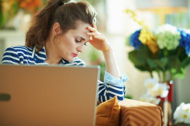 Şık, orta yaşlı, mavi bluzlu ve çizgili ceketli bir kadın güneşli bir günde modern evde dizüstü bilgisayarla divanda oturuyor..