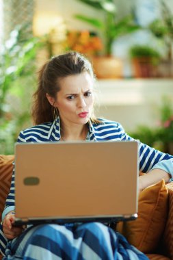 Şok olmuş 40 yaşındaki mavi bluzlu ve çizgili ceketli kadın güneşli bir günde modern evde dizüstü bilgisayarla divanda oturuyor..