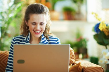 Gülümseyen, mavi bluzlu ve çizgili ceketli modern kadın güneşli bir günde modern evde dizüstü bilgisayarla divanda oturuyor..