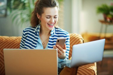 Güneşli bir günde modern evde dizüstü bilgisayarlar, akıllı telefonlar kullanarak mavi bluzlu ve çizgili ceketli gülümseyen genç bir ev hanımı..