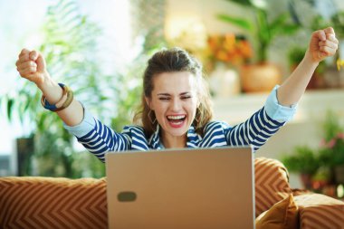 Mavi bluzlu, çizgili ceketli ve dizüstü bilgisayarı olan orta yaşlı bir kadın kanepeye oturmuş güneşli bir günde modern evde eğleniyor..