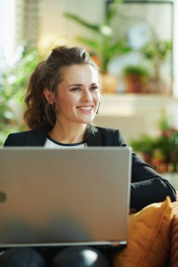 Mutlu modern 40 yaşındaki beyaz bluz ve siyah ceketli kadın güneşli bir günde modern evde dizüstü bilgisayarın uzaklığına bakıyor ve kanepede otururken internette sörf yapıyor..