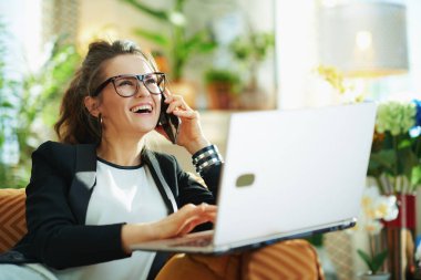 Mutlu, orta yaşlı, beyaz bluzlu, siyah ceketli, dizüstü bilgisayarlı, güneşli bir günde modern oturma odasında cep telefonu kullanan bir ev hanımı..