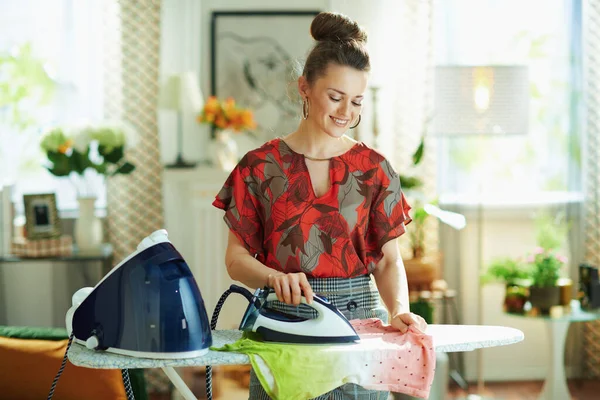 smiling young housewife in red blouse and grey pencil skirt ironing with steam generator on ironing board in the modern house in sunny day.