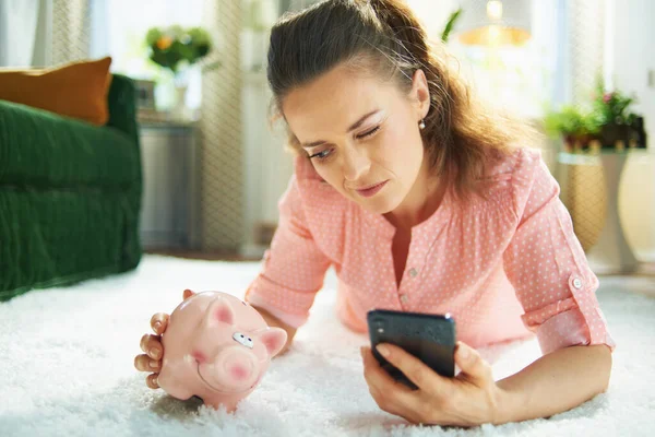 modern 40 years old woman in blouse and white pants with piggy bank searching for carpet cleaning service on a smartphone while laying on white carpet in the modern living room in sunny day.