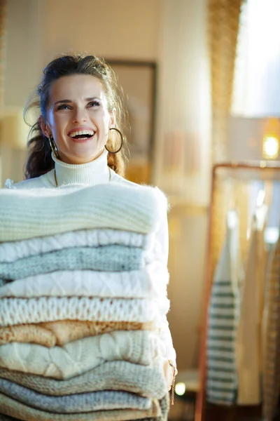 白いセーターとスカートの若い女性が笑顔で晴れ冬の日に現代のリビングルームで大量のセーターを持って — ストック写真