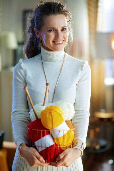 smiling trendy female in white sweater and skirt holding basket with knitting yarn and needles in the modern house in sunny winter day.