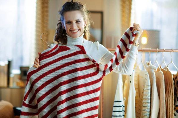若いです40歳の女性で白いセーターとスカートで現代の家で晴れた冬の日に銅の服ラックにぶら下がっているセーターを選択 — ストック写真