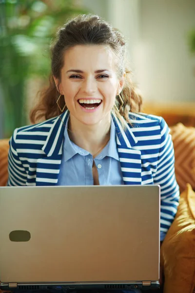 阳光明媚的现代家中 穿着蓝色衬衫和条纹夹克 头戴笔记本电脑坐在沙发上的快乐年轻女子的画像 — 图库照片