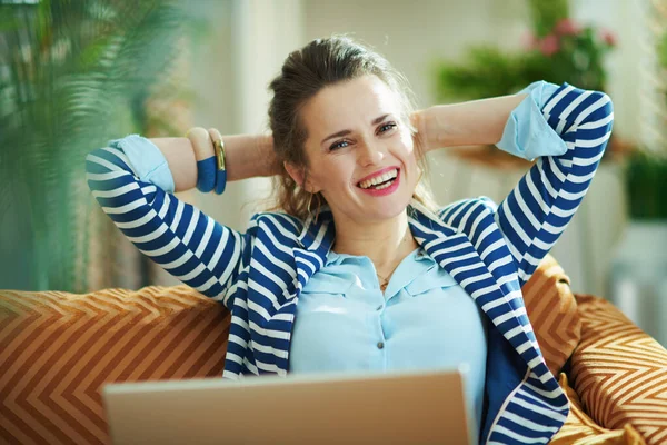 在阳光明媚的日子里 穿着蓝色衬衫和条纹夹克 头戴笔记本电脑 坐在沙发上的中年妇女笑了 — 图库照片