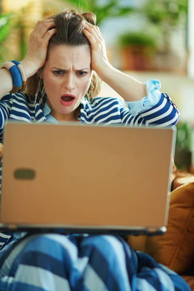 Şok olmuş mavi bluzlu ve çizgili ceketli şık ev hanımı güneşli bir günde modern evde dizüstü bilgisayarla divanda oturuyor..