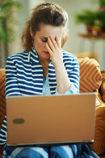 stressed elegant woman in blue blouse and striped jacket sitting on couch with laptop at modern home in sunny day.