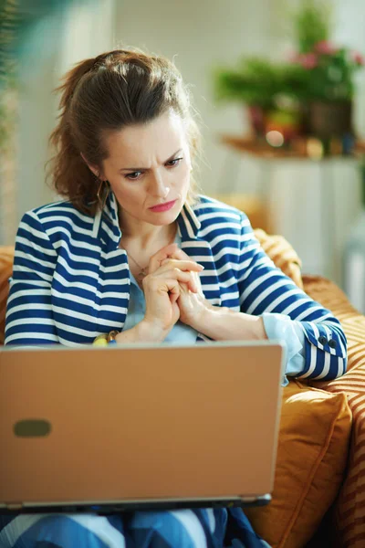 阳光明媚的今天 穿着蓝色衬衫 穿着条纹夹克 头戴笔记本电脑 坐在沙发上的中年不幸的家庭主妇 — 图库照片
