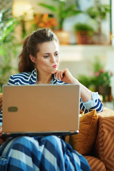 阳光明媚的日子里 穿着蓝色衬衫和条纹夹克 头脑清醒的中年妇女坐在沙发上 还带着笔记本电脑坐在现代房子里 — 图库照片