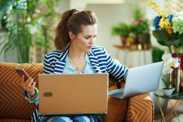 在阳光灿烂的日子里 穿着蓝色衬衫和条纹夹克的中年时髦家庭主妇们在现代家中使用着不同的设备 笔记本电脑 智能手机 — 图库照片