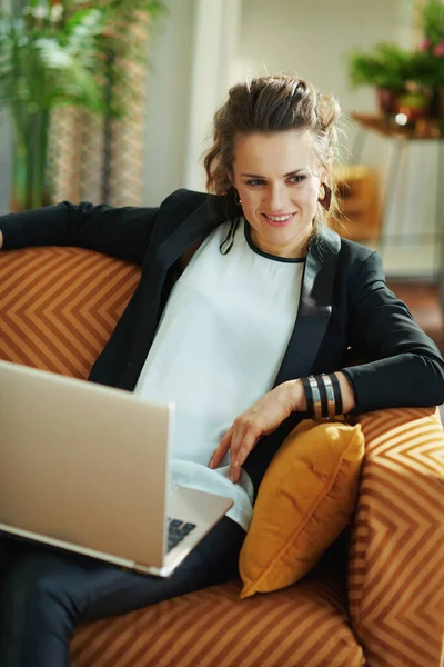 在现代客厅里 穿着白衬衫和黑色夹克的中年女性在阳光灿烂的日子里坐在沙发上阅读笔记本电脑上的文字 — 图库照片