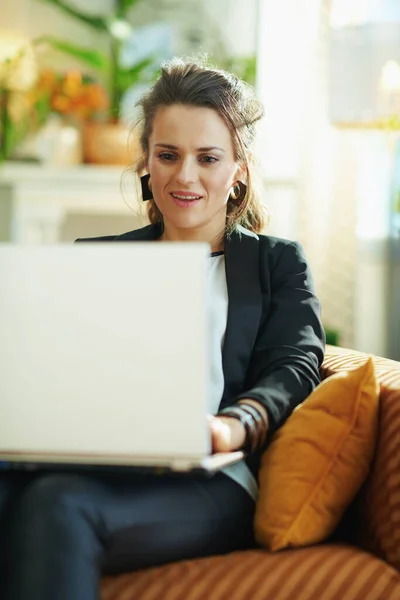 在现代客厅里 穿着白衬衫和黑色夹克的时尚女性在阳光灿烂的日子里坐在沙发上阅读笔记本电脑上的文字 — 图库照片
