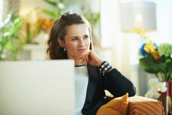 pensive trendy woman in white blouse and black jacket at modern home in sunny day looked aside while reading text on a laptop.