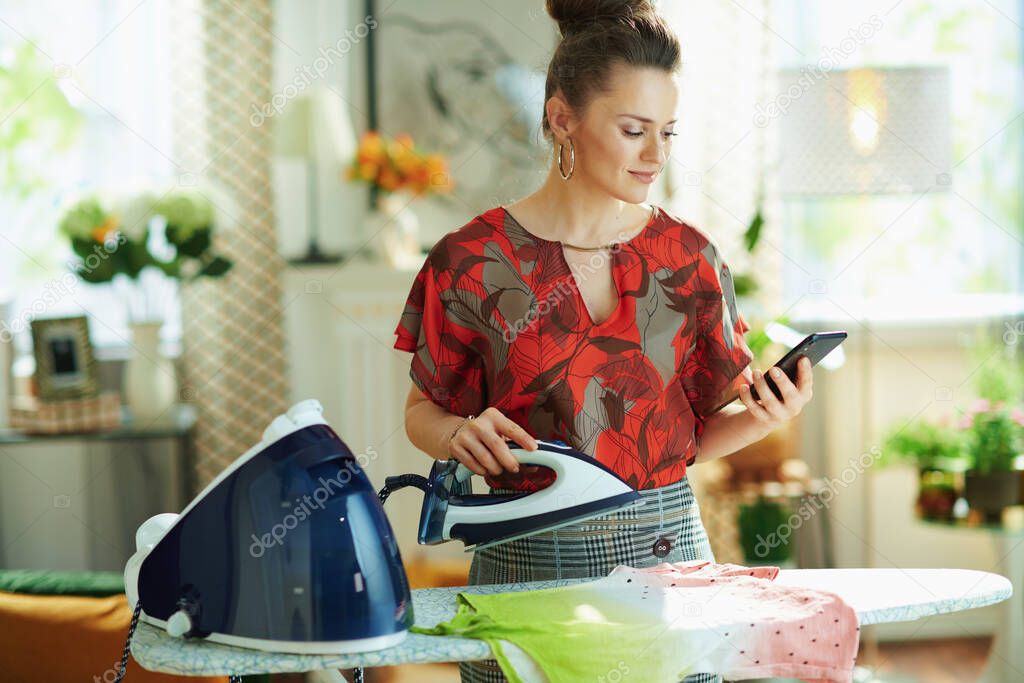 elegant woman in red blouse and grey pencil skirt ironing with steam generator on ironing board and checking how to iron correctly on internet on a smartphone in the modern house in sunny day.