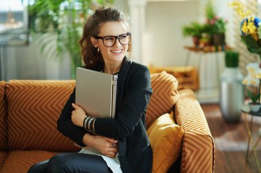 Mutlu, 40 yaşında, beyaz bluzlu ve siyah ceketli bir kadın güneşli bir günde modern oturma odasında kanepede oturuyor ve dizüstü bilgisayarına sarılıyor..