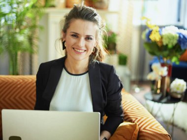 Güneşli bir günde modern evde beyaz bluz ve siyah ceketli mutlu orta yaşlı ev hanımı divanda otururken dizüstü bilgisayarda yazıyor..