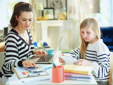 Anne, çocuk ev ödevlerini yaparken güneşli bir günde modern oturma odasında çalışıyor..