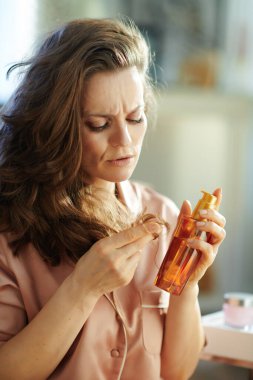 Uzun kahverengi saçlı, uzun pijamalı genç bir kadın saç yağı şişesi tutuyor ve güneşli kış günlerinde modern evde saç uçlarını kontrol ediyor..