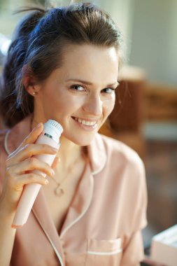 Mutlu modern 40 yıllık ev hanımı pijamalarıyla güneşli kış gününde modern oturma odasında elektrikli yüz temizleme fırçası kullanıyor..