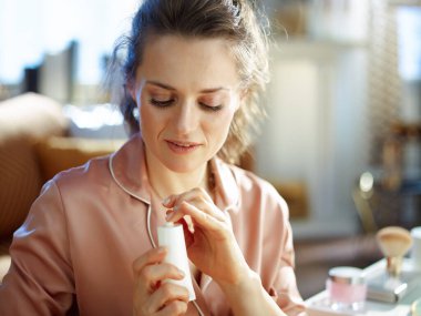 Güneşli kış gününde modern evde beyaz kozmetik tüplü, pijamalı 40 yaşında gülümseyen bir kadın..