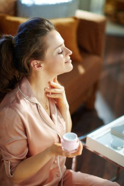 Pijamalı, pembe kozmetik kavanozlu mutlu orta yaşlı ev hanımı güneşli kış gününde modern oturma odasına boyun kremi sürüyor..