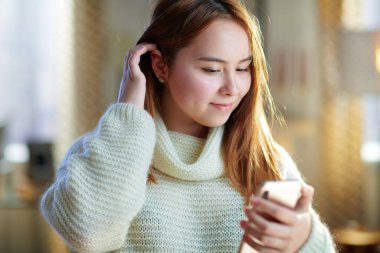 Güneşli kış gününde modern oturma odasında beyaz kazaklı mutlu modern genç kız sosyal medya profil ekranına bakıyor..