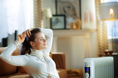 Güneşli kış gününde modern evde beyaz kazak ve etek giyen rahat stil sahibi bir kadın kanepenin ve beyaz elektrikli kaloriferin yanında oturuyor..