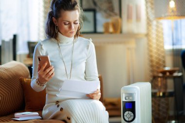 Beyaz kazaklı ve etekli orta yaşlı bir kadının güneşli kış günlerinde modern evde beyaz elektrikli kaloriferin yanında oturduğundan kuşkuluyum..