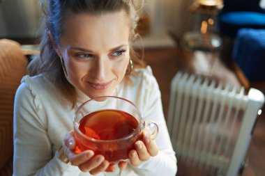 Modern modern orta yaşlı ev hanımının portresi güneşli kış gününde, beyaz elektrikli kaloriferin yanındaki kanepede oturup sıcak çay içiyor..