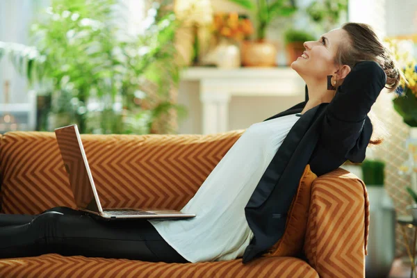 在阳光明媚的日子里 身穿白衬衫和黑色夹克 头戴笔记本电脑的40岁女人坐在现代客厅的沙发上 轻松而时尚 — 图库照片