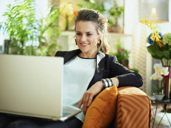 白いブラウスに黒いジャケットを着た現代の主婦がソファに座っている間にノートパソコンでウェブサイトを使って — ストック写真