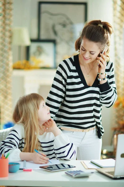 Anne akıllı bir telefonla konuşuyor, çalışıyor ve çocuğa ev ödevlerinde yardım ediyor. Güneşli bir günde modern oturma odasında..