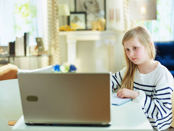 young child in striped sweater in the modern living room in sunny day checking website on a laptop while sitting at the table.