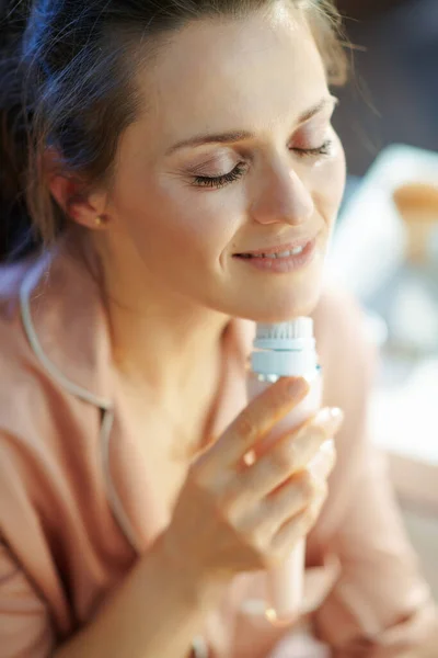 relaxed modern woman in pajamas in the modern living room in sunny winter day using electric facial cleansing brush.